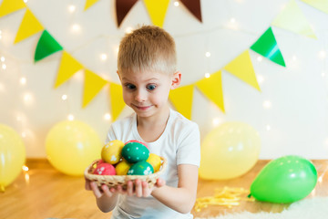 blond, blue-eyed cute smiling boy in a white shirt, the age of 6 years, with easter eggs in a room on the background of yellow design, sitting, studio