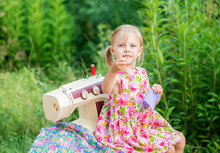 Little Blonde Smiling Lovely Child 4 Years Old (girl) In Cotton Dress Sewing Needle (hand) Of The Purple Cloth On A Background Of A Sewing Machine With A Cotton Cloth Beautiful Outdoors