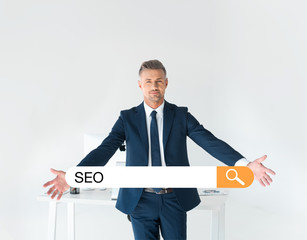 Wall Mural - handsome businessman standing with open arms near table with computer isolated on grey with seo in search bar