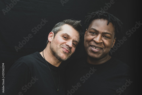 Fotografía An interracial gay couple pose before the camera to have their pictures taken - black man and mixed race man with stubble - on a black background - with copy space