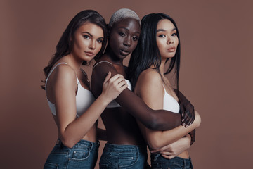 Simply stunning. Three attractive young women looking at camera while standing against brown background