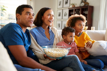 Wall Mural - Young family sitting together on the sofa in their living room watching TV and eating popcorn, side view