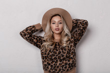 Attractive Young Woman In An Elegant Trendy Beige Hat In A Trendy Leopard Sweater With Curly Blond Hair With Natural Makeup With Sexy Lips Near A White Wall In The Studio. Stylish Girl.