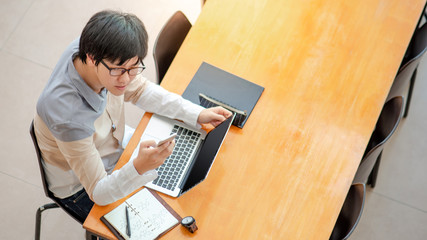 Wall Mural - Young Asian man university student using smartphone while working with laptop computer and notebook on wooden table in the college. Campus lifestyle in education building. Shot from top view