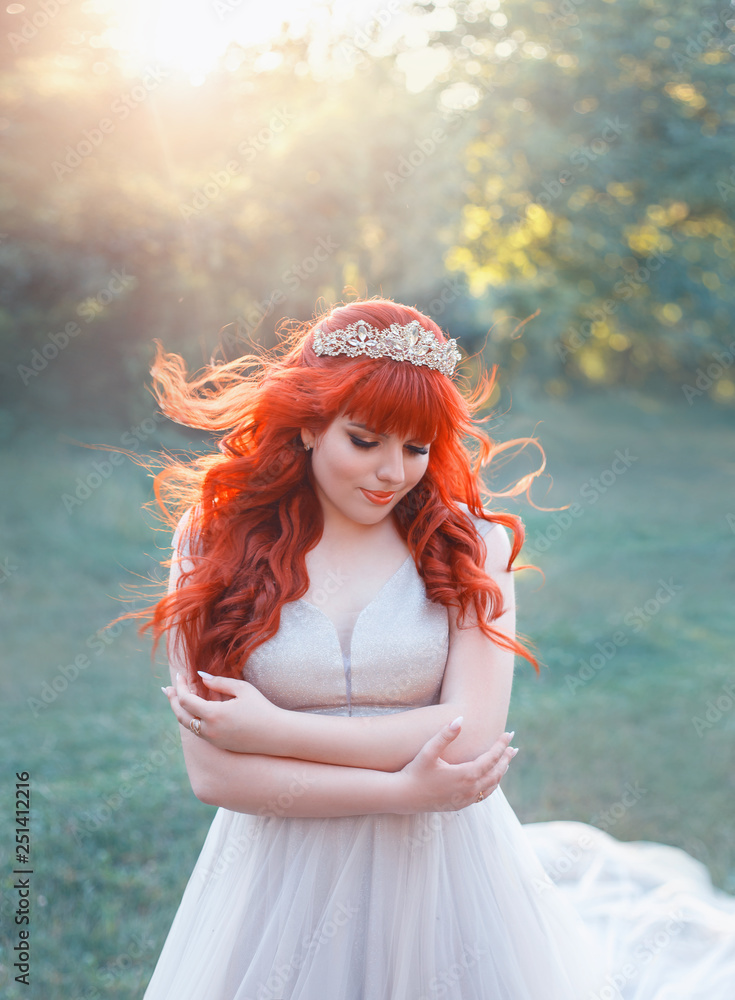 attractive girl with bright red hair and bangs stands alone on green glade,  hugging herself and