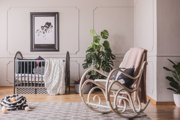 Rocking chair and poster in grey child's bedroom interior with plant and blanket on cradle. Real photo