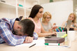 © Zoriana - View of male student tired student sleeping on his books, during lecture at university. Group of students sitting at table, reading book on blur background in class.