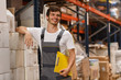 © Nestor - Cheerful worker wearing uniform and white t shirt, holding yellow clipboard. Handsome man smiling, standing and leaning on white boxes in warehouse. Concept of entrepot and commercial industry.