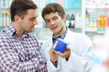 Pharmacist in white coat holding blue pack and showing it to male customer. Good looking man in checked shirt looking at it. Men standing in drugstore. Concept of pharmaceutical industry.