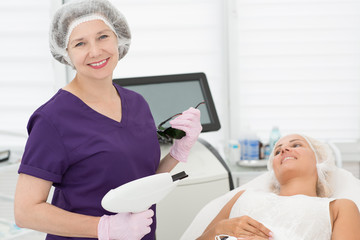 Wall Mural - Qualified dermatologist in process of doing ultrasound procedure in cosmetology office. Female beautician in uniform and disposable hat keeping apparatus in hands, looking at camera and posing.