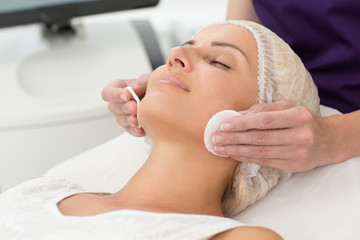 Wall Mural - Closeup of face of female client lying on couch and enjoying process during cosmetic procedure in cosmetology office. Hands of professional beautician wiping skin with cotton disks. Skin care concept.