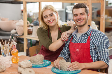 Charming couple in aprons mastering new favourite craft in workshop and enjoying production process. Man and woman sitting together, looking at camera and smiling while sculpting earthenware.