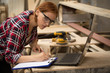 © Zoriana - Busy and serious woman with ginger hair working at carpentry. Professional craftswoman writing on sheet of paper. Lumbers, wood, notebook and special equipment on background.
