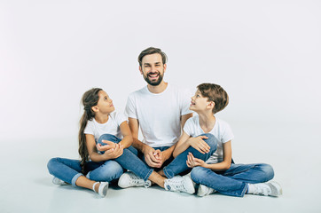 Wall Mural - Handsome young father, attractive son and cute daughter are sitting together on the floor and hugging each other with happy and smiling faces. Best family