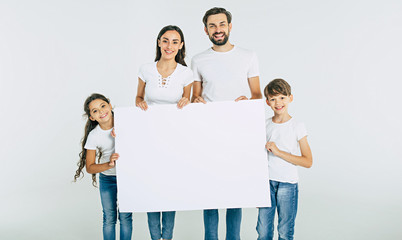 beautiful smiling family in white t-shirts hold in hands the big blank banner and looking on camera
