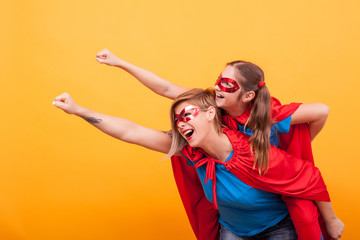 Beautiful mother and her cute little girl playing together dressed like superheros over yellow background.