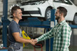 Smiling worker of car service in uniform and satisfied client shaking hands. Happy man in checkered shirt thankful mechanic for repaired automobile. Concept of dealing and agreement.