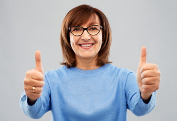 gesture, vision and old people concept - portrait of smiling senior woman in glasses showing thumbs up over grey background