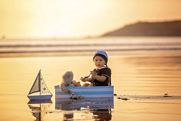 Cute baby child, sweet boy, playing with boat, teddy bear and fishes on sunset at the edge of the ocean