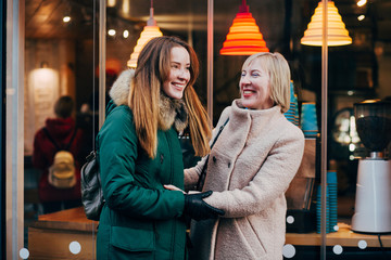 Beautiful mature mother and her adult daughter are talking, smiling, walking together outdoor in winter city street. Bonding, different generationsconnect, family concept.