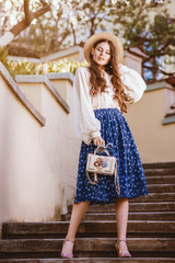 Outdoor full body portrait of young beautiful fashionable happy woman posing in spring city. Model wearing straw hat, stylish white blouse, blue skirt, holding small bag. Sunny day light