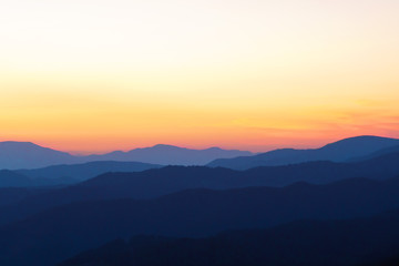 Poster - Panorama of the peaks of beautiful mountains covered with trees at sunset on a sunny day in summer. Orange glow on the sky after full sunset. Dark silhouettes of mountains. Solar reflection