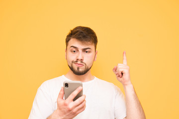 Wall Mural - Emotional man with a beard holds a smartphone in his hands, showing his finger up in a blank space. Surprised adult in a white T-shirt uses a smartphone and shows a finger on copyspace