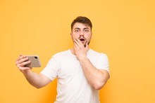Shocked Man With A Beard Standing On A Yellow Background With A Smartphone , Looks At The Camera With Astonishment. Amazed Gamer With A Smartphone In His Hands Is Isolated On A Yellow Background.