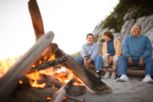 Mature Couple Sitting With Their Teenage Grandson Beside A Bonfire On A Beach.