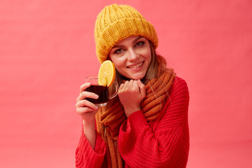 Girl in a red sweater and knitted hat holding a cup of hot mulled wine with an orange slice. Red background. Studio