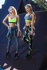 Two nice and positive sport fit girls posing with dumbbells in skatepark. Looking at camera and smiling. Girls relaxing after intensive workout in city park. Healthy lifestyle