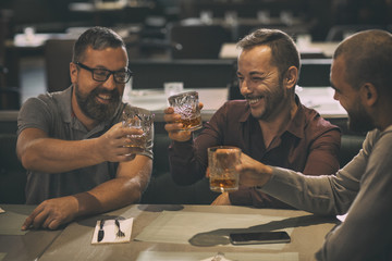 Wall Mural - Cheerful smiling men holding glasses with alcohol beverages and laughing. Three friends spending time together and having fun in bar. Men drinking scotch, whiskey or brandy.