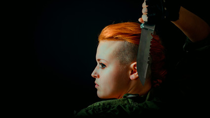 Young woman with red hair stand back in military uniform, keep a knife in hand. Horizontal photo