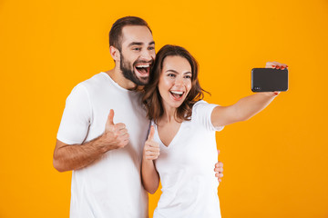 Poster - Portrait of two happy people man and woman taking selfie photo on smartphone, isolated over yellow background