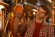 © Nestor - Concentrated and serious inspector of beer factory holding glass of beer in hand and looking at it. Handsome man in checked shirt inspecting and checking quality of beverage.