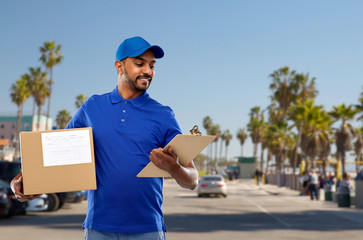 Wall Mural - mail service and shipment concept - happy indian delivery man with parcel box and clipboard in blue uniform over venice beach background in california