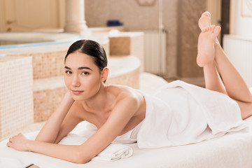 attractive asian woman in towel lying on massage table and looking at camera