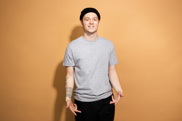 Smiling guy dressed in a gray shirt, black jeans and black hat with tattoo on his hand is posing on the beige background in the studio