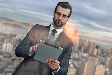 Wall Mural - Using modern technologies. Handsome bearded businessman in suit using digital tablet while standing outdoors with cityscape on the background