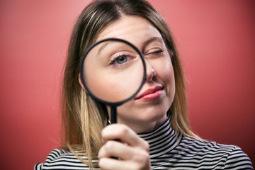 Cheerful young woman looking through magnifying glass at the camera over pink background.