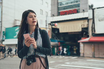 Wall Mural - vintage asian girl traveler carrying slr camera using cellphone searching online map app finding direction standing on zebra cross in osaka japan. Translation on wall building text 