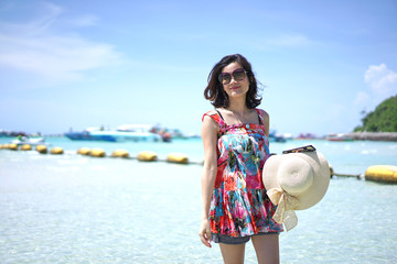 Wall Mural - Portrait of young Asian woman standing on the beach with beautiful sea in background. She wearing sunglasses and holding her hat with smile. She is feeling happy on her vacation.