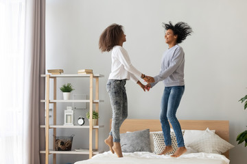 Wall Mural - Black mother and adolescent daughter holding hands jumping on bed