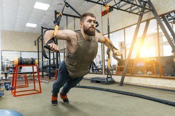 Wall Mural - Muscular bearded man dressed in military weighted armored vest doing exercises using straps systems in the gym. Sport, training, bodybuilding and healthy lifestyle concept.