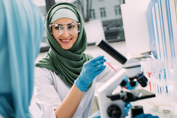 smiling female muslim scientist in goggles and hijab during experiment in chemical laboratory