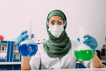 Wall Mural - surprised female muslim scientist looking at camera and holding flasks during experiment in chemical laboratory