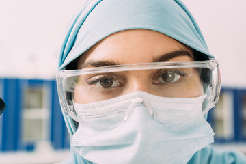 Wall Mural - close up of female muslim scientist in medical mask, goggles and hijab looking at camera