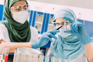 Wall Mural - female muslim scientists in medical masks holding glass ampoule during experiment in chemical laboratory