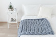 Bed with white linen and grey knitted woolen merino chunky blanket. Light stylish cozy scandinavian bedroom interior. Blanket of thick yarn. Geometric glass florarium with plants on bedside table.