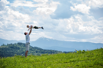 Happy father throws up a little son against a background of green forest, mountains and sky with clouds. Paternity friendship